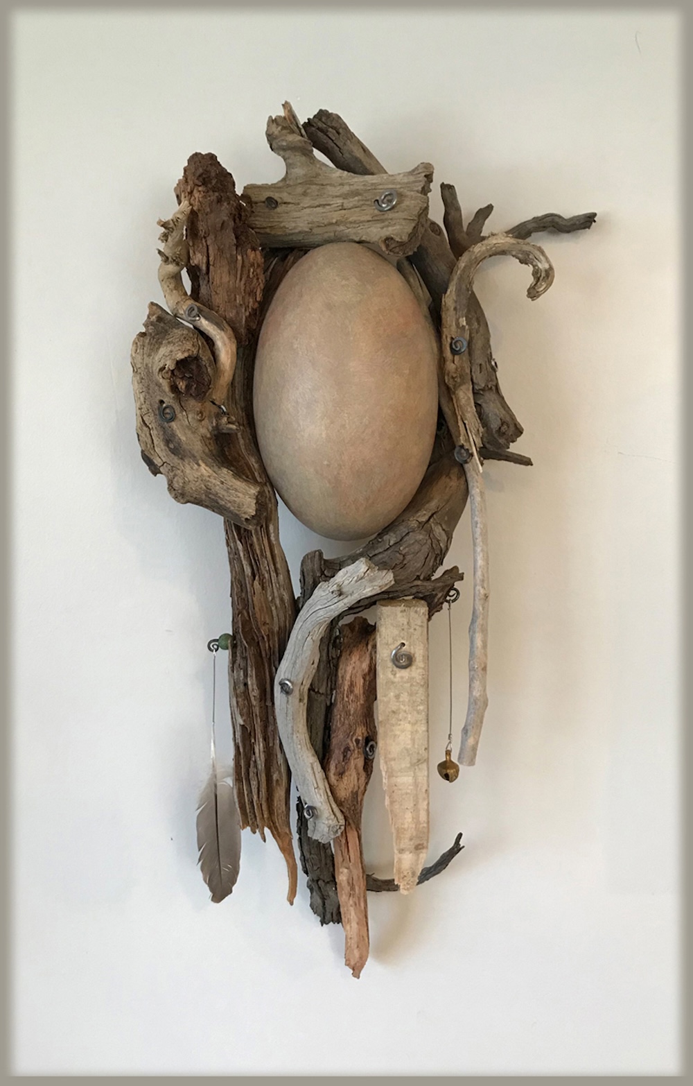 Monochromatic wall sculpture made of found wood, a feather, and held together with spiral-shaped metal pieces. A ceramic egg shape is nestled in the center.