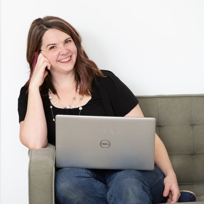 Woman (Meg Casebolt) with long brown hair on a sofa leaning on her right arm with a computer in her lap.