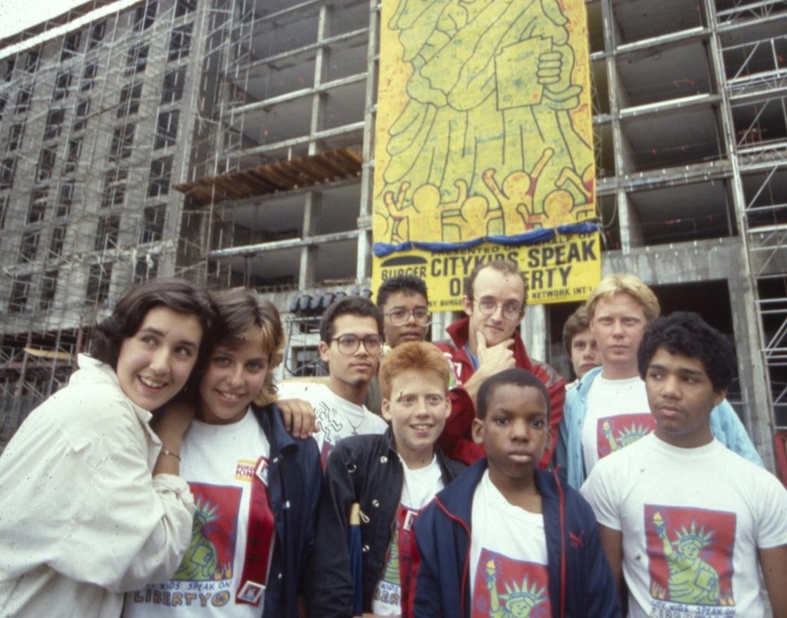 Keith Haring and CityKids participants standing beneath the large Liberty banner in 1986